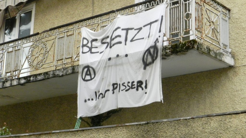 Ansicht des Balkons des Hauses. Hier ist ein Transparent mit einem Anarchie-A und dem Symbol der Hausbesetzer_innen angebracht. Aufschrift "Besetzt! … ihr Pisser!"