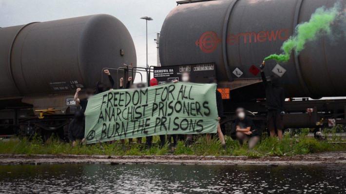Eine Gruppe verpixelter Leute mit einem grünen Transparent mit der Aufschrift "Freedom for all anarchist prisoners. Burn the prisons".