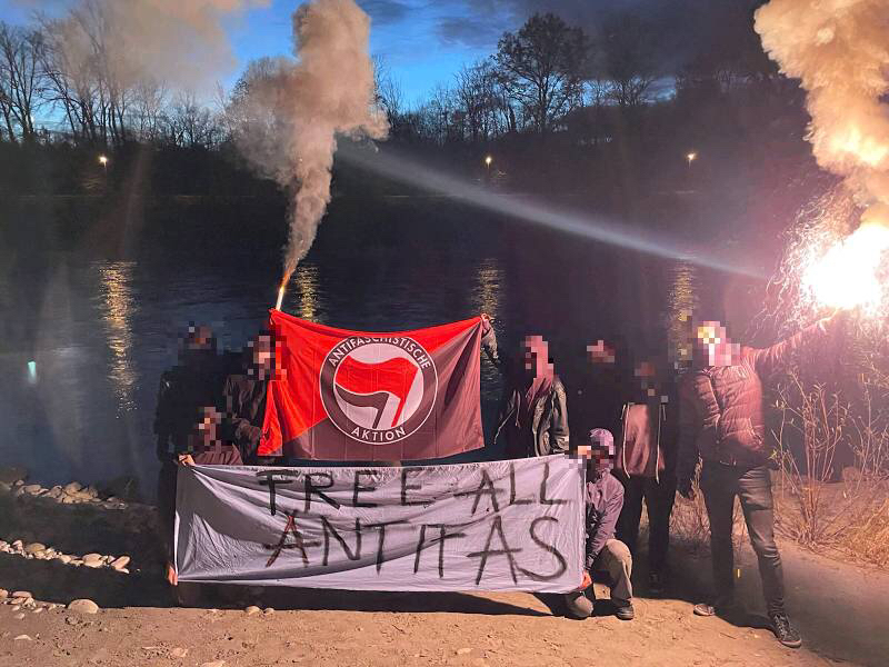 Mehrere verpixelte Personen mit einem Transparent mit der Aufschrift "Free all Antifas", einer schwarz-roten Antifa-Fahne und Pyrotechnik.